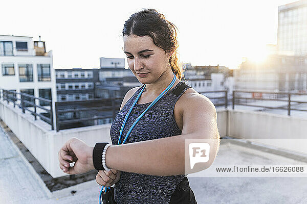Young woman with jump rope checking time while standing on terrace at sunset