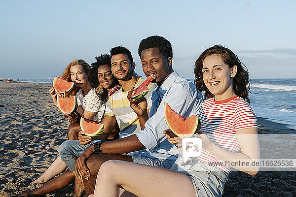 Smiling friends eating watermelon while sitting on beach during sunny day