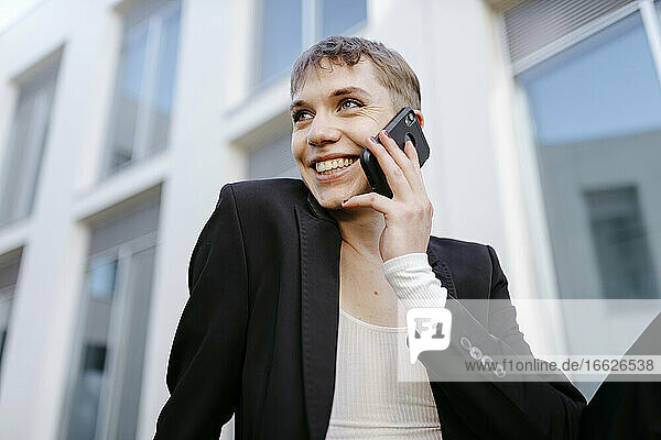 Nahaufnahme eines lächelnden trans jungen Mannes  der über ein Mobiltelefon spricht  während er an einem Gebäude sitzt