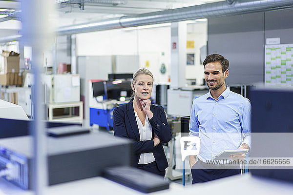 Lächelnde Berufskollegen diskutieren  während sie sich Maschinen in einer beleuchteten Fabrik ansehen