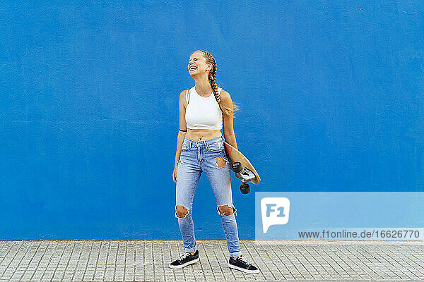 Fröhliche Frau mit Skateboard auf dem Fußweg vor einer blauen Wand