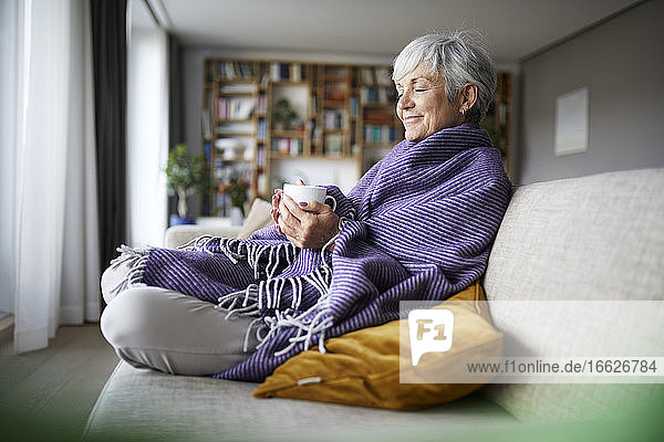 Nachdenkliche ältere Frau mit Decke  die eine Kaffeetasse hält  während sie zu Hause auf dem Sofa sitzt