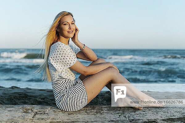 Schöne junge Frau sitzt auf einem Baumstamm am Strand während eines sonnigen Tages