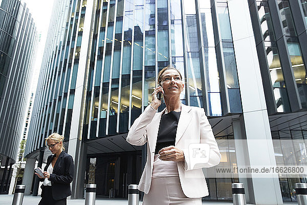 Weibliche Fachkraft  die mit einem Mobiltelefon spricht  während sie neben einer Geschäftsfrau vor einem modernen Bürogebäude steht