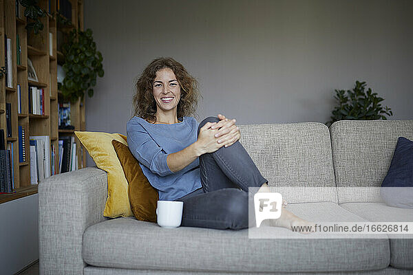 Lächelnde Frau trinkt Kaffee  während sie zu Hause auf dem Sofa sitzt