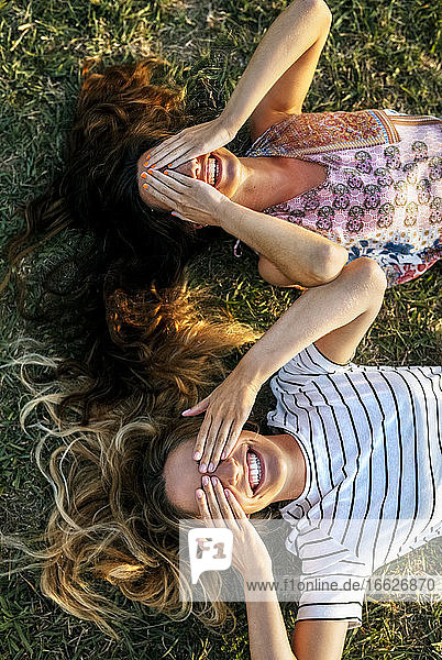 Fröhliche junge Frauen bedecken ihre Augen mit den Händen  während sie auf einem Feld liegen