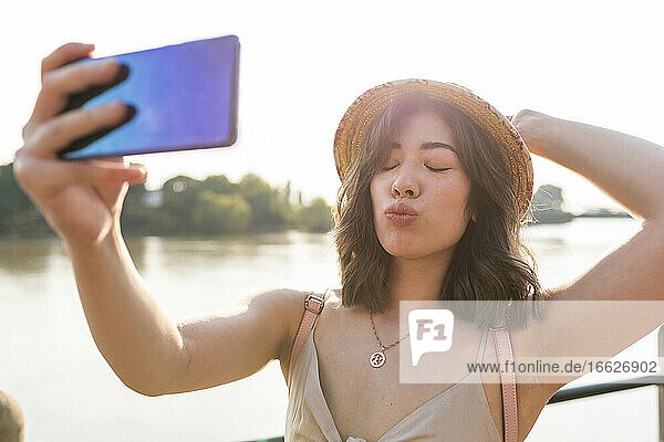 Beautiful woman grimacing while taking selfie on smart phone standing against river