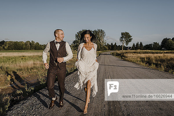 Happy bride and bridegroom running on road during sunny day