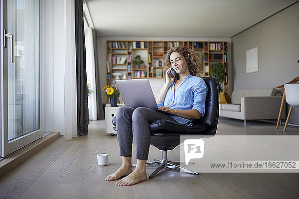 Frau  die mit einem Mobiltelefon spricht  während sie zu Hause einen Laptop benutzt