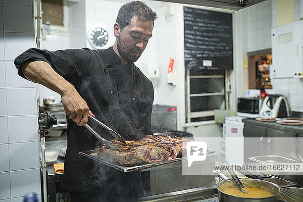 Chef using serving tongs while arranging grilled meat in tray at kitchen