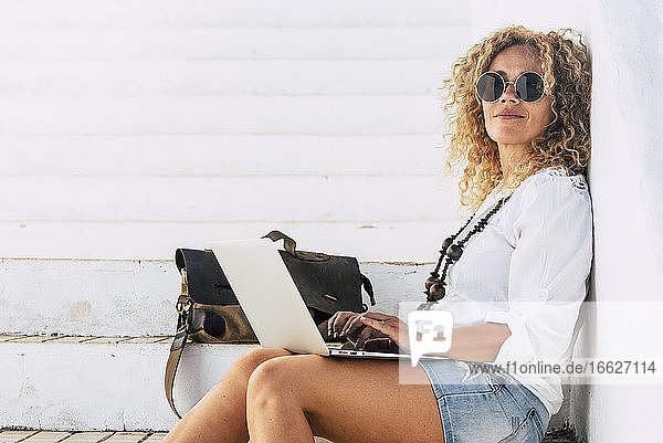 Selbstbewusste Frau mit Sonnenbrille  die einen Laptop benutzt  während sie auf einer Treppe sitzt