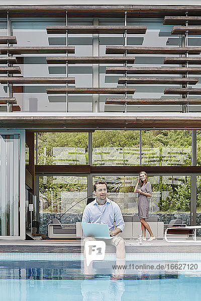 Mann arbeitet am Laptop und schaut weg  während er an einem modernen Haus sitzt