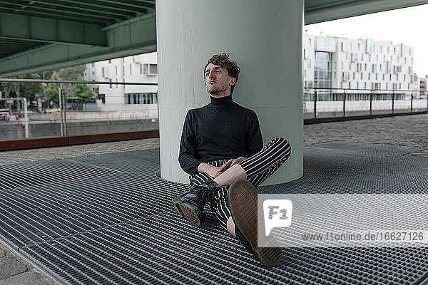 Thoughtful young man looking away while sitting by column on footpath