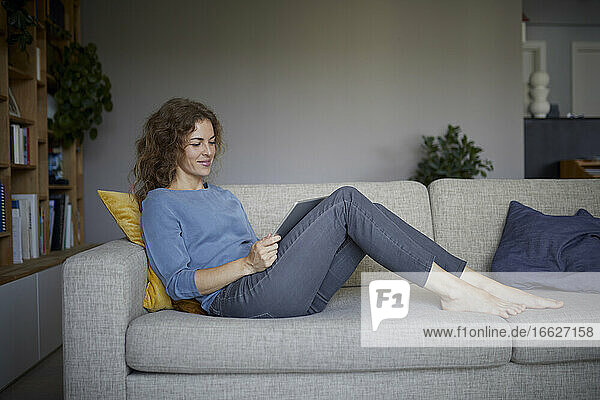 Lächelnde Frau  die ein digitales Tablet benutzt  während sie zu Hause auf dem Sofa sitzt