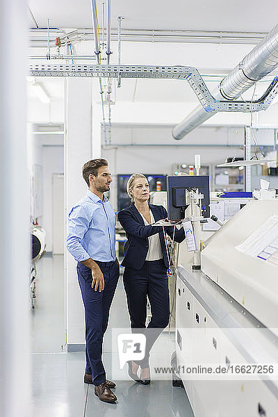 Männliche und weibliche Kollegen diskutieren  während sie auf einen Computer an einer Maschine in einer beleuchteten Fabrik schauen