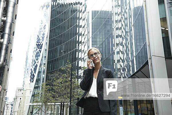 Blond female entrepreneur talking on phone while standing against office building at downtown district