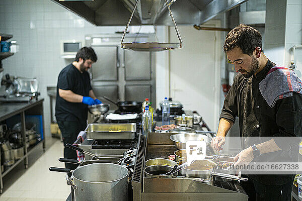 Chef standing by kitchen counter with coworker cooking in background at kitchen