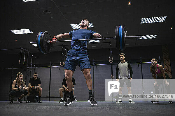 Adaptive athlete jumping while lifting barbell with people cheering in background at gym