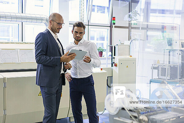 Confident male colleagues discussing over digital tablet while standing against machinery at factory