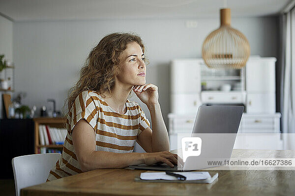 Nachdenkliche Frau mit Kopf in den Händen  die einen Laptop benutzt  während sie zu Hause am Tisch sitzt