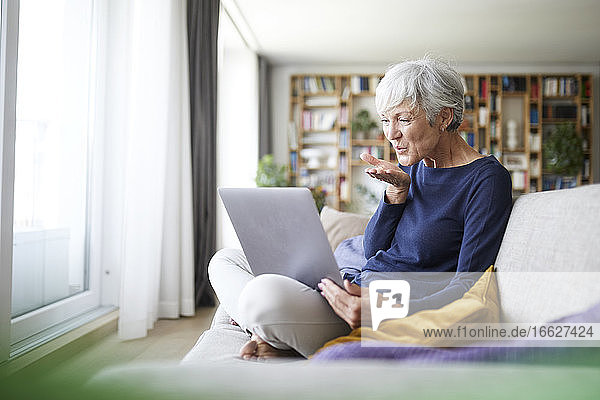 Ältere Frau gibt fliegenden Kuss auf Videoanruf  während sie zu Hause sitzt