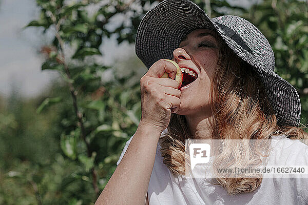 Close-up of happy woman wearing hat eating apple in orchard