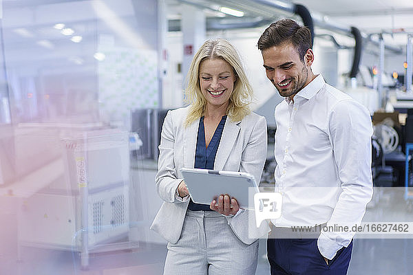 Lächelnde blonde Geschäftsfrau  die mit einem jungen männlichen Kollegen in einer Fabrik über ein digitales Tablet diskutiert