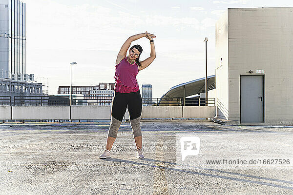 Junge Frau mit erhobenen Armen  die auf einer Terrasse vor dem Himmel stehend Sport treibt