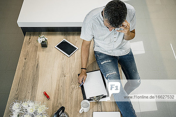 Male entrepreneur talking through smart phone while sitting with clipboard on desk in office