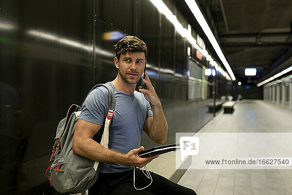 Gut aussehender junger Mann  der telefoniert und ein digitales Tablet hält  während er auf eine beleuchtete U-Bahn blickt