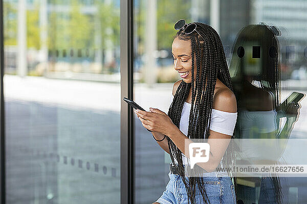 Woman using phone while leaning on glass wall in city