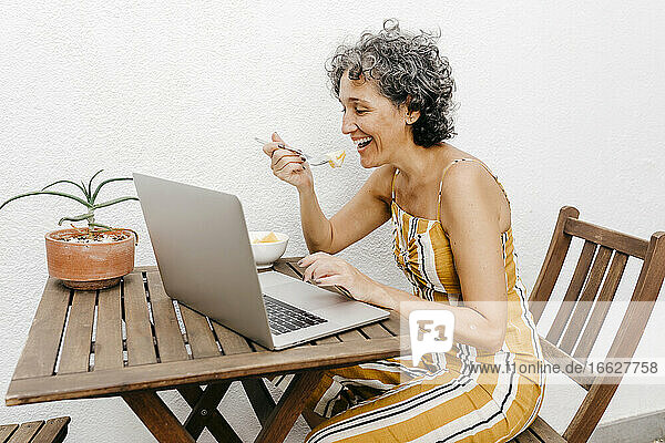 Lächelnde reife Frau  die an einem Tisch an einer weißen Wand im Hinterhof isst und dabei einen Laptop benutzt