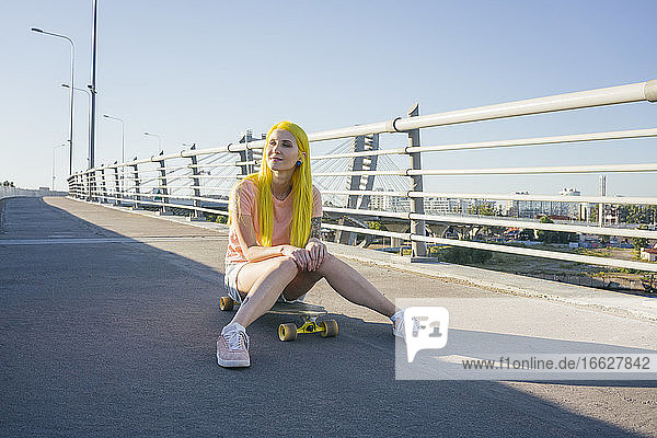 Junge Frau schaut weg  während sie auf einer Brücke gegen den klaren Himmel an einem sonnigen Tag sitzt