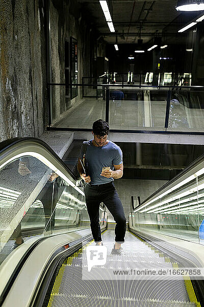 Männlicher Pendler  der ein Mobiltelefon benutzt  während er auf einer Rolltreppe in der U-Bahn steht