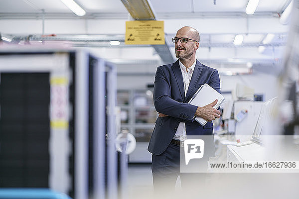 Lächelnder nachdenklicher männlicher Fachmann  der ein digitales Tablet hält und in eine helle Fabrik schaut