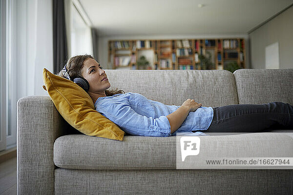 Mittlere erwachsene Frau mit Kopfhörern  die Musik hört  während sie zu Hause auf dem Sofa liegt