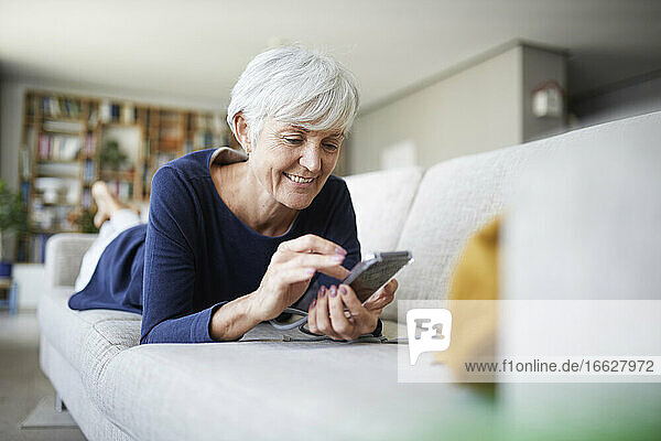 Ältere Frau benutzt ein Smartphone  während sie zu Hause auf dem Sofa liegt