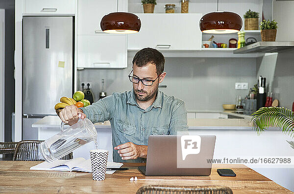 Geschäftsmann gießt Wasser aus einem Krug in ein Glas  während er mit seinem Laptop zu Hause sitzt