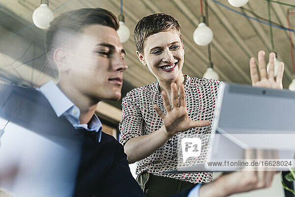 Lächelnde Geschäftsfrau  die über ein digitales Tablet gestikuliert  das von einem männlichen Kollegen im Büro gehalten wird