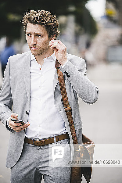 Businessman with mobile phone wearing in-ear headphones while standing in city