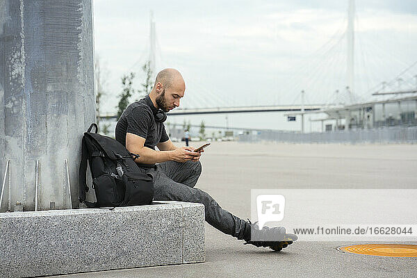 Junger Mann in Rollschuhen  der ein Mobiltelefon benutzt  während er auf einem Sitz in der Stadt sitzt