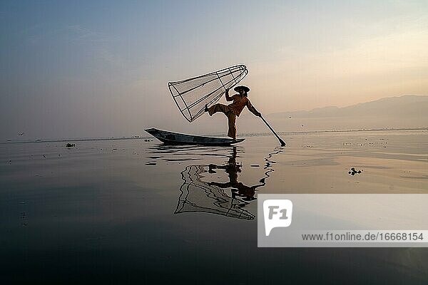 Traditional fisherman posing standing on his small boat with reflection in front of sunrise Lake Inle Myanmar Asia
