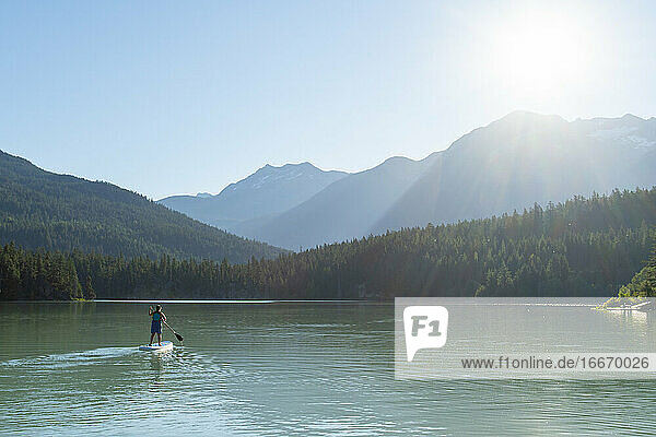 Rückenansicht einer anonymen Frau auf einem SUP-Board auf einem friedlichen See in der Nähe von Bergen und Wäldern an einem sonnigen Tag im Hochland von British Columbia  Kanada