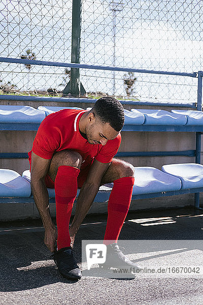 African American male soccer player sitting and fixing boots on bench