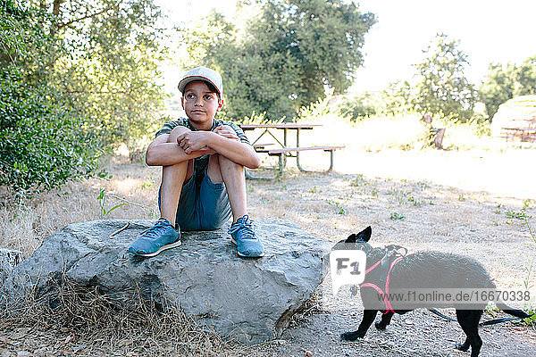 Boy sits on a large rock daydreaming with his dog nearby