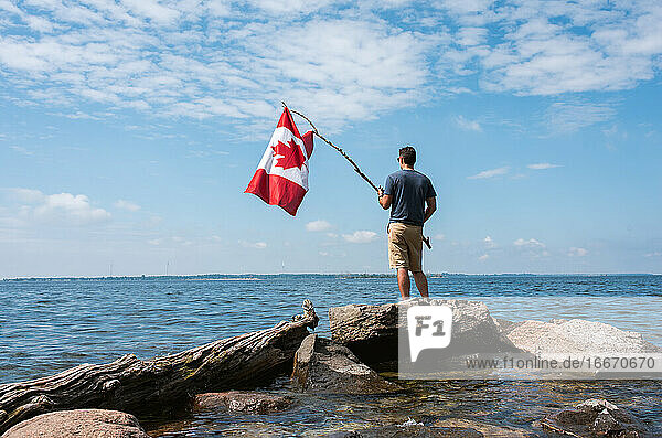 Mann hält die kanadische Flagge am felsigen Ufer eines Sees an einem Sommertag.