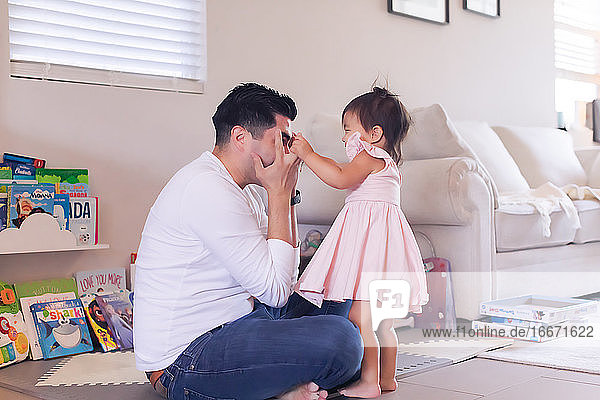 Dad sitting on the floor playing peekaboo with baby daughter.
