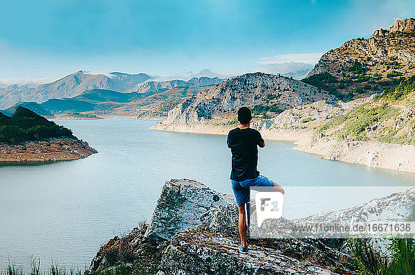 Junger Mann balanciert in Baumpose auf einem Felsen in einem sonnigen  abgelegenen Tal