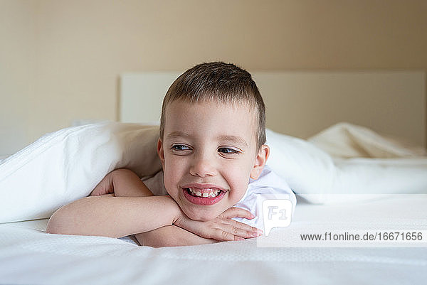 Adorable smiling kid lying down on bed under blanket  looking camera