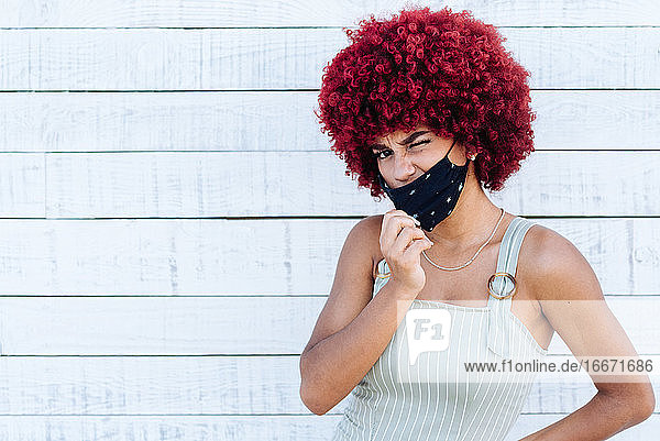 Latino-Frau mit rotem Afro-Haar und Maske  die an einer weißen Wand steht.